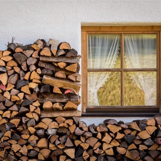 A window with curtains behind which there are stacks of wood. The logs are neatly arranged and create a rustic atmosphere.