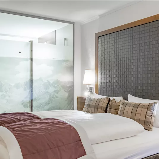 A modern bedroom with a large bed and elegant decor. A glass wall showcases a picturesque mountain landscape.