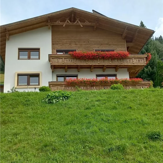 Ein schönes Haus auf einer Wiese mit Balkonen voller Blumen. Im Hintergrund sind Bäume und ein klarer Himmel zu sehen.