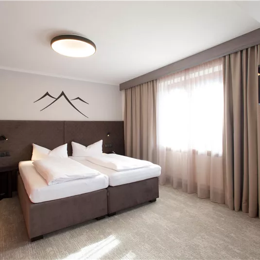 A modern hotel room with two single beds and a television. Bright curtains let in light and the walls are simply decorated.