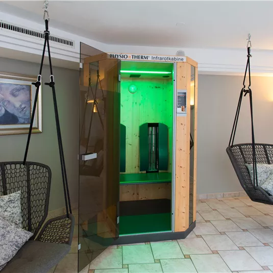 A modern wellness room with an infrared cabin and two rocking chairs. The lighting is kept in a soft green.