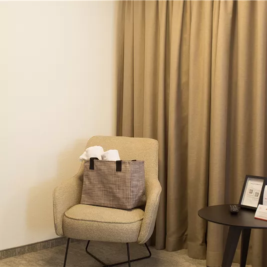 A cozy armchair with a pocket stands next to a small table in a bright room. Curtains and a mirror complement the modern decor.