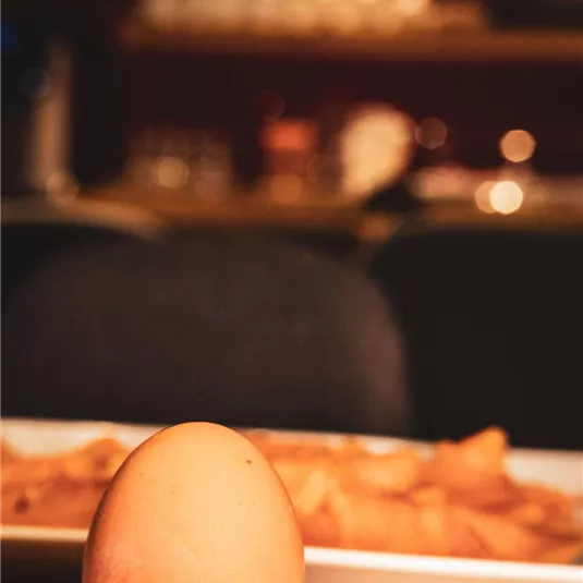 A boiled egg on a small plate with a spoon. In the background, some delicious dishes and a cozy atmosphere can be seen.
