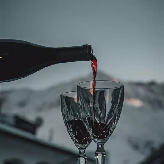 A bottle of red wine is poured into two glasses. Snow-covered mountains can be seen in the background.
