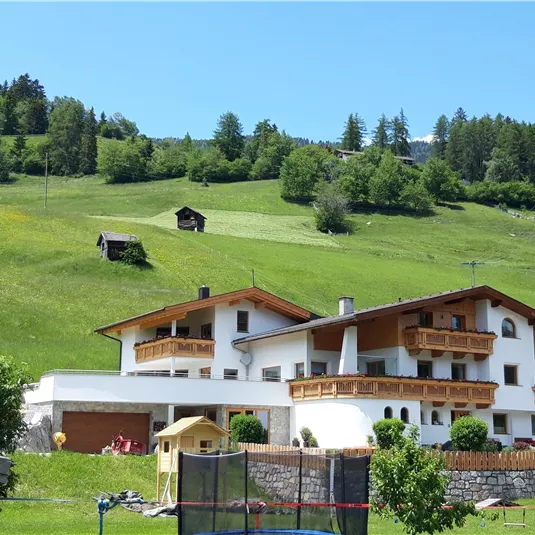 Ein großes, modernes Haus mit Holzbalkonen steht in einer grünen Hügellandschaft. Im Hintergrund sind Bäume und eine kleine Hütte sichtbar.