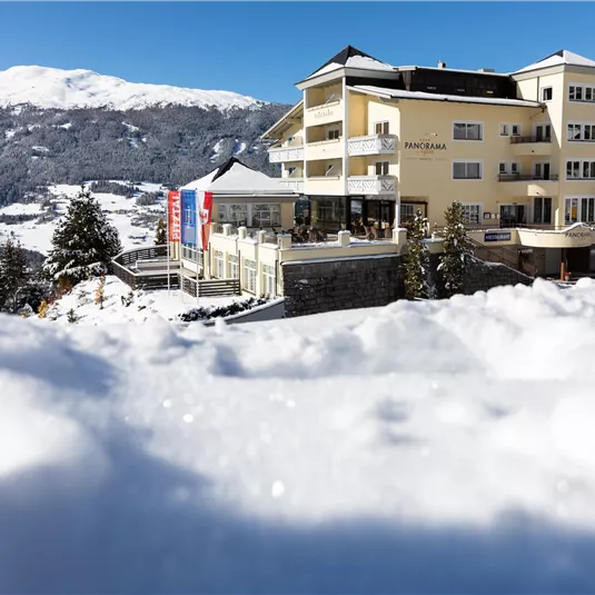 A cozy hotel in a wintry landscape. Snow covers the ground while the mountains in the background shine.