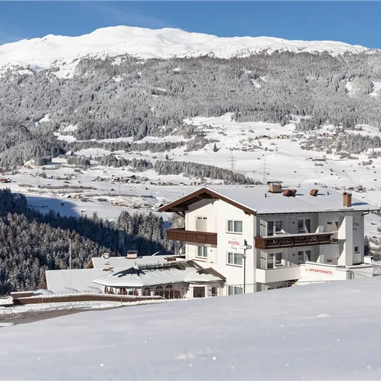 A snow-covered mountainous landscape with a white building in the foreground. The scenery is clear and sunny, surrounded by tall, snowy mountains.