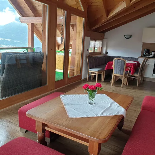 A cozy living room with a red sofa, a wooden table, and a view of the mountains. The large window front provides plenty of daylight and an inviting atmosphere.