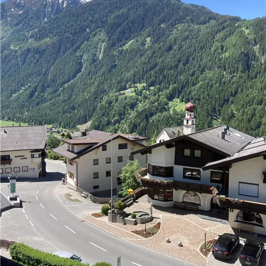 Eine malerische Berglandschaft mit grünen Hügeln und schneebedeckten Gipfeln. Im Vordergrund sind charmante Gebäude und eine ruhige Straße zu sehen.