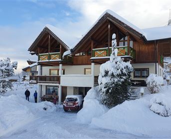 A beautiful house in the snow with a red car in front. The surroundings are surrounded by snow-covered trees and mountains.