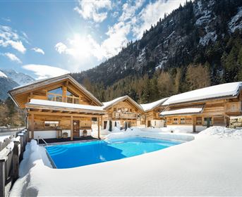 A beautiful outdoor area with a blue swimming pool, surrounded by snow-covered cabins. In the background, majestic mountains and a clear, sunny sky can be seen.