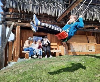 Ein Kind schwingt auf einer Schaukel vor einem Holzhaus. Im Hintergrund sitzen mehrere Personen und genießen die Zeit im Freien.
