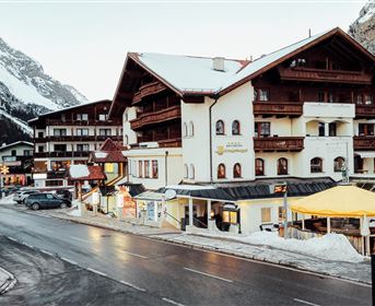 Ein charmantes Bergdorf mit traditionellen Chalets und schneebedeckten Dächern. Die Straße ist leicht befahren und im Hintergrund sind majestätische Berge sichtbar.