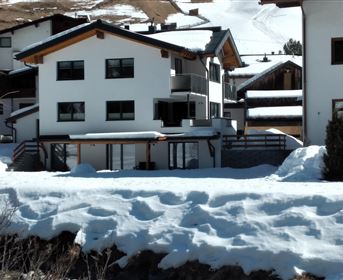A modern house in the snow with a large balcony. In the background, additional buildings and snow-covered hills can be seen.