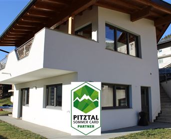 A modern house with large windows and a balcony. In the foreground, a green area can be seen.