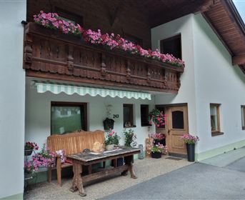 An inviting house with blooming balcony plants and a wooden bench in front of the door. The facade is bright and well-maintained, with colorful flowers in pots.