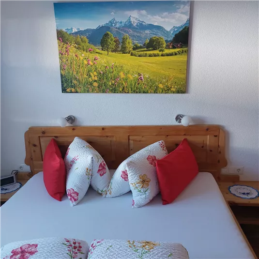 A cozy bedroom with a wooden bed and colorful cushions. On the wall, there is a picture of a picturesque landscape with mountains and meadows.