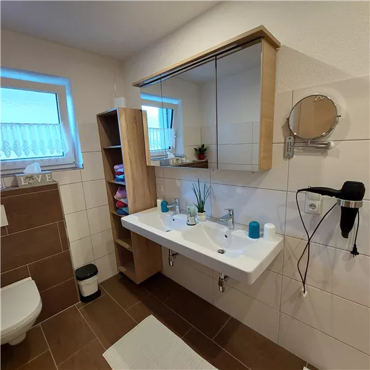 Ein modernes Badezimmer mit einem Doppelwaschbecken und einem Spiegel. Es ist hell und gemütlich, mit Holzregalen und einer Pflanze.