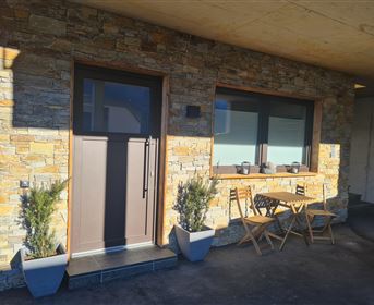 Eine moderne Haustür mit einer Steinverkleidung und großen Fenstern. Vor der Tür stehen zwei Stühle und ein kleiner Tisch in einer warmen, einladenden Atmosphäre.