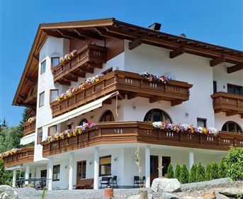 Ein gemütliches Alpenhaus mit Holzbalkonen und bunten Blumen. Es steht in einer klaren und sonnigen Umgebung.