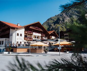 Ein traditionelles Alpenhotel mit mehreren Etagen und Balkonen. Im Vordergrund befinden sich Sonnenschirme und die beeindruckende Berglandschaft im Hintergrund.