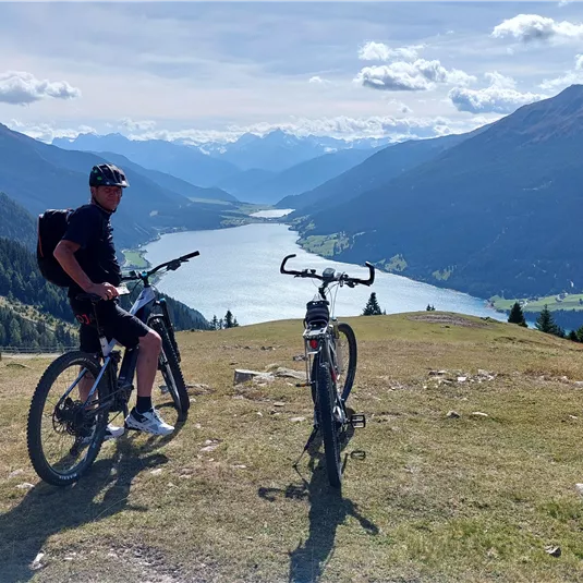 A cyclist stands in a meadow overlooking a beautiful lake and surrounding mountains. The landscape is sunny and offers a stunning view.