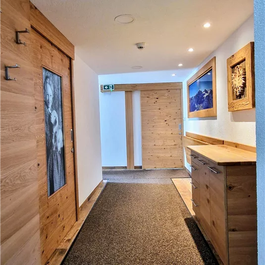 Ein heller Flur mit Holzverkleidung und großen Türen. An den Wänden hängen Bilder, und der Boden ist mit einem Teppich ausgelegt.