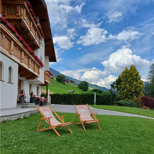 Eine grüne Wiese mit zwei Liegestühlen steht vor einem charmanten Alpenhaus. Der Himmel ist blau mit einigen Wolken und die Umgebung ist malerisch.