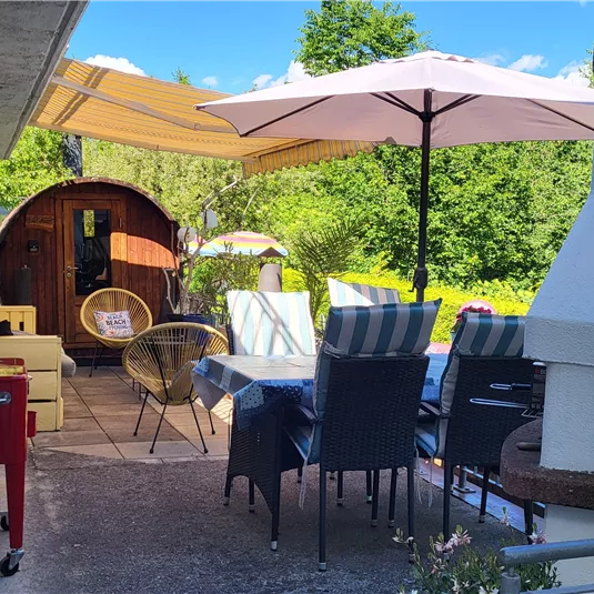 Eine gemütliche Terrasse mit einem Esstisch und Stühlen. Im Hintergrund sind üppige Bäume und Sonnenschirme zu sehen.
