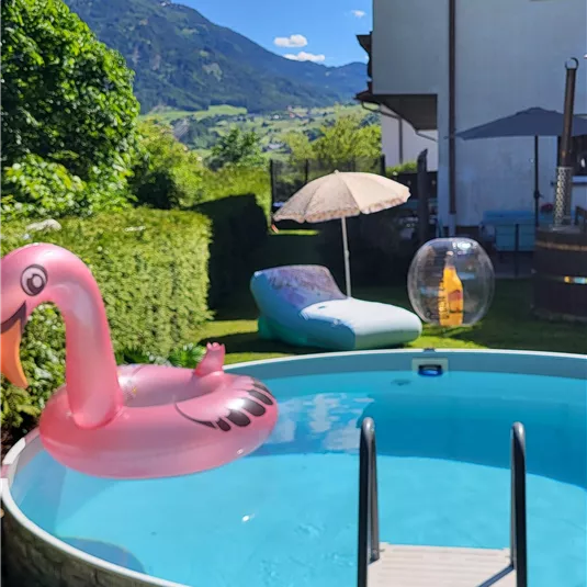 Ein schöner Pool im Garten mit einem rosa Flamingo und sonnigem Wetter. Im Hintergrund sind grüne Berge und ein schattiger Sonnenschirm zu sehen.