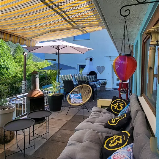 Ein gemütlicher Außenbereich mit einem Sofa, Sesseln und bunten Kissen. Ein Sonnenschirm spendet Schatten und verschiedene Pflanzen sorgen für eine angenehme Atmosphäre.