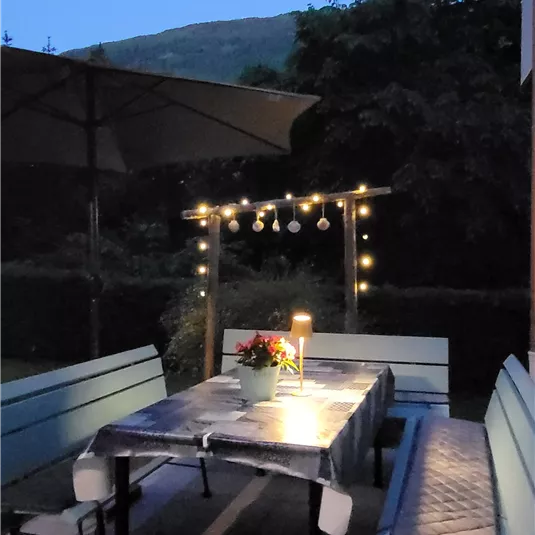 Ein gemütlicher Außenbereich mit einem Tisch und Bänken. Lichter und eine Sonnenschirm schaffen eine einladende Atmosphäre in der Abenddämmerung.