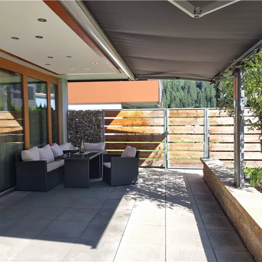 A modern terrace with cozy seating furniture and sun protection. The area is bright and inviting, surrounded by plants and a fence.