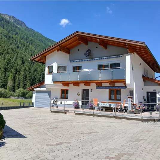 A modern house in the mountains with a large outdoor area. The surroundings are surrounded by green forests and hills.