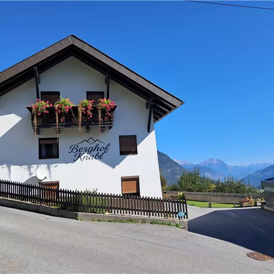 A picturesque house with flowers on the balcony, surrounded by mountains. The sky is blue and clear.