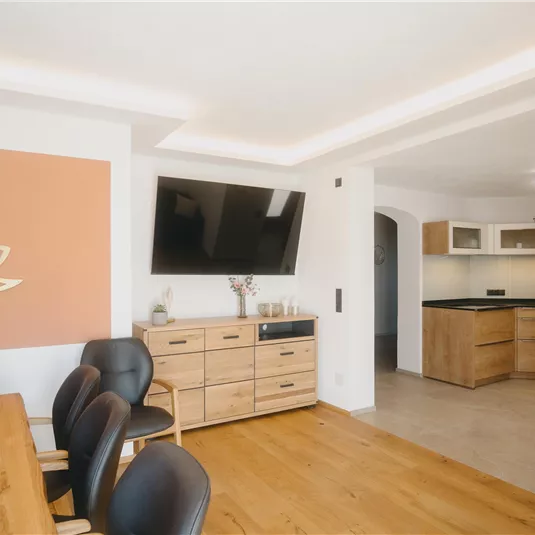 A modern dining room with a wooden table and black chairs. The kitchen is open and has light furniture as well as a large television on the wall.