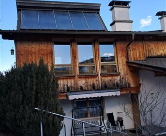 A charming wooden house with solar panels on the roof. The garden is covered in snow and surrounded by a small walkway.