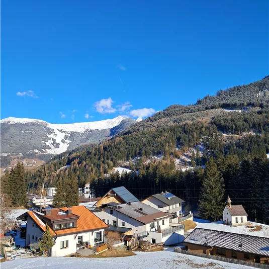 Eine malerische Berglandschaft mit schneebedeckten Bergen und einem klaren blauen Himmel. Im Vordergrund sieht man ein kleines Dorf mit charmanten Häusern.