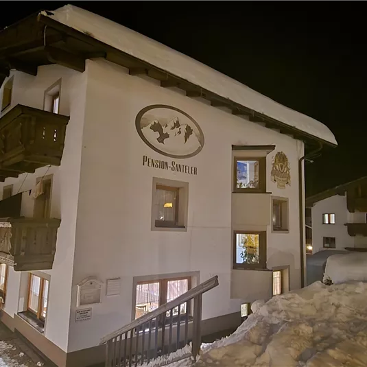 Ein gemütliches Gästehaus im Schnee mit warmem Schein in den Fenstern. Die Szene zeigt eine winterliche Nachtstimmung.