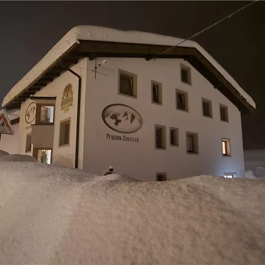 Ein verschneites Gebäude bei Nacht mit einer ruhigen Atmosphäre. Der Schnee bedeckt den Boden und das Licht erhellt die Fassade.