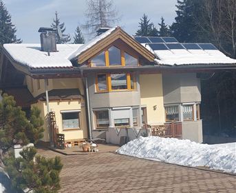 Ein modernes Haus mit Solarpanelen auf dem Dach steht in einem schneebedeckten Gelände. Im Hintergrund sind hohe Bäume sichtbar.