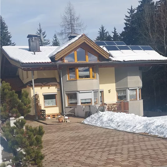 Ein modernes Haus mit Solarpanelen auf dem Dach steht in einem schneebedeckten Gelände. Im Hintergrund sind hohe Bäume sichtbar.
