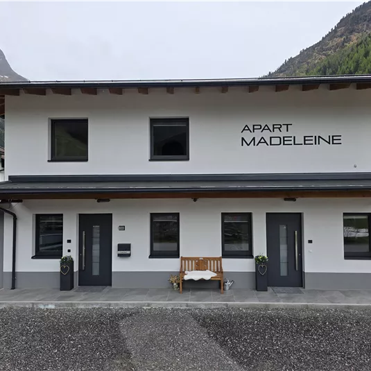 Ein modernes Gebäude mit der Aufschrift "Apart Madeleine". Es hat eine helle Fassade und liegt in einer bergigen Umgebung.