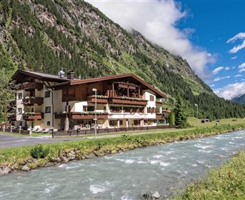 A charming building by the riverside, surrounded by green meadows and impressive mountains. The sky is clear and the scenery is picturesque.