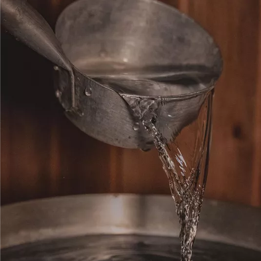 A metal spoon is pouring water into a bowl. In the background, a wooden paneling can be seen.