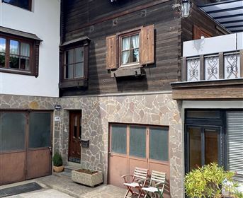 Ein traditionelles altes Haus mit Holzfassade und großen Fenstern. Vor der Tür stehen bunte Gartenstühle und ein kleiner Topf mit Pflanzen.