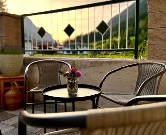 Ein gemütlicher Balkon mit zwei Stühlen und einem kleinen Tisch. Auf dem Tisch steht eine Vase mit bunten Blumen und die Berge im Hintergrund sind gut sichtbar.