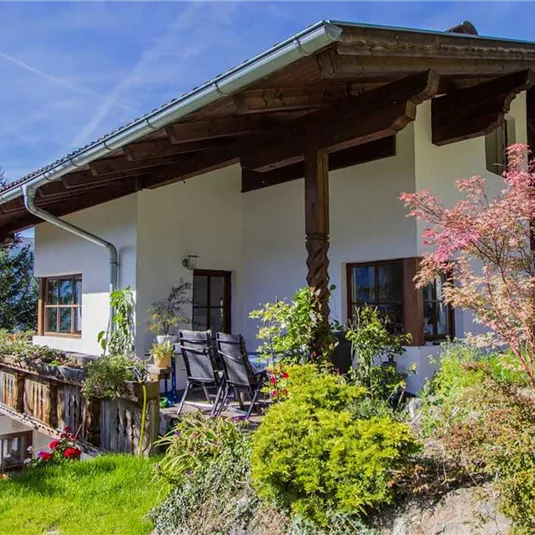 A pretty house in a green garden, surrounded by trees. The terrace is equipped with plants and a cozy seating area.