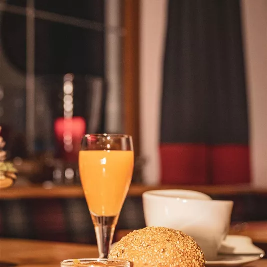 A beautiful breakfast table with a roll, a small portion of jam, and a glass of orange juice. In the background, a cup of coffee can be seen.