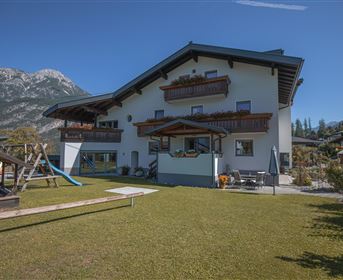 Ein gut gepflegtes Haus mit mehreren Balkonen in einer grünen Umgebung. Im Vordergrund gibt es einen Spielplatz und die Berge im Hintergrund sind sichtbar.